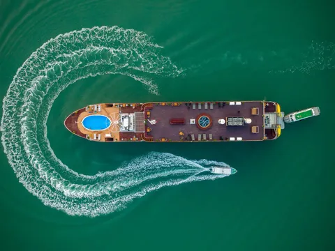 Aerial view of a cruise boat with a blue pool on the front deck moving through emerald green water, with two smaller boats creating white wake trails in an S-shaped pattern around it.