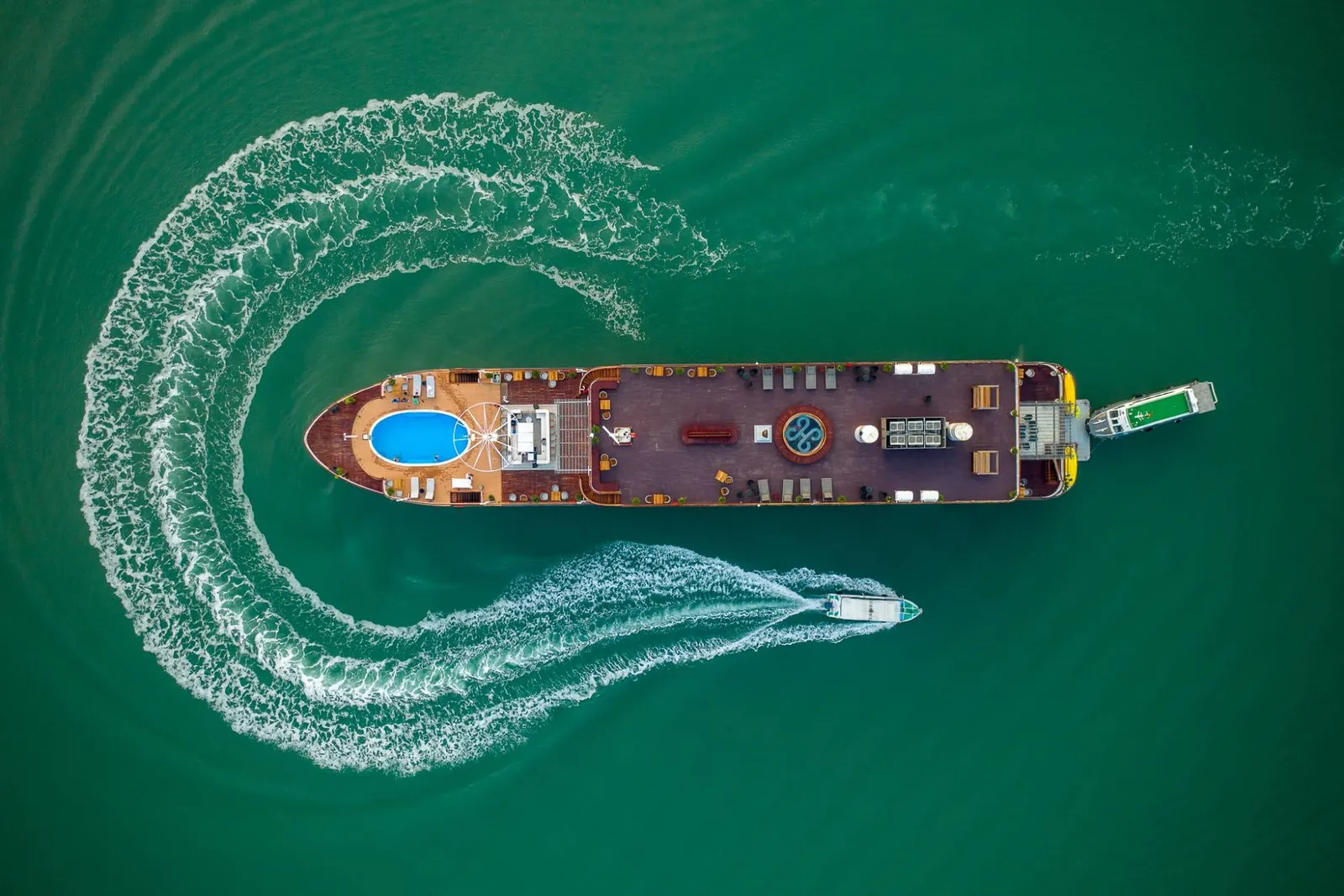 Aerial view of a cruise boat with a blue pool on the front deck moving through emerald green water, with two smaller boats creating white wake trails in an S-shaped pattern around it.