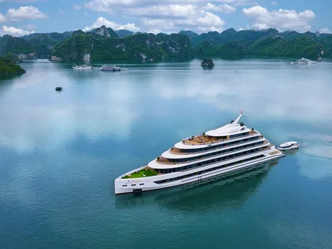Aerial view of a large white multi-deck cruise ship with a small tender boat beside it on calm turquoise water, surrounded by green limestone karst islands and a few distant boats under a partly cloudy sky.