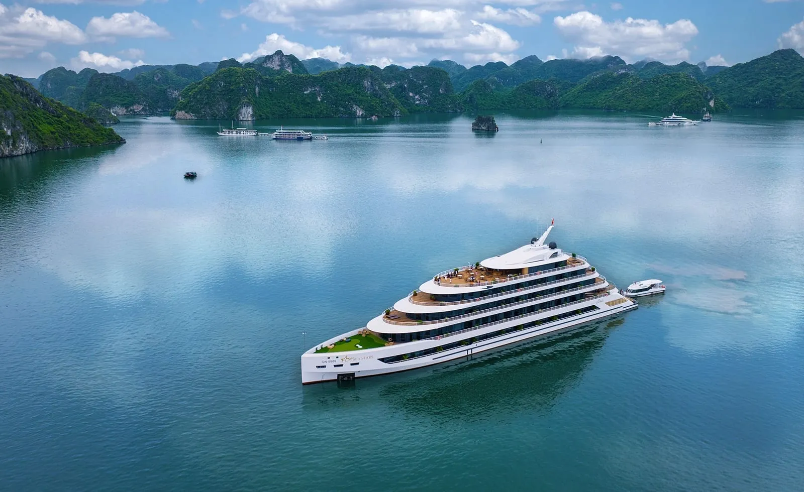 Aerial view of a large white multi-deck cruise ship with a small tender boat beside it on calm turquoise water, surrounded by green limestone karst islands and a few distant boats under a partly cloudy sky.