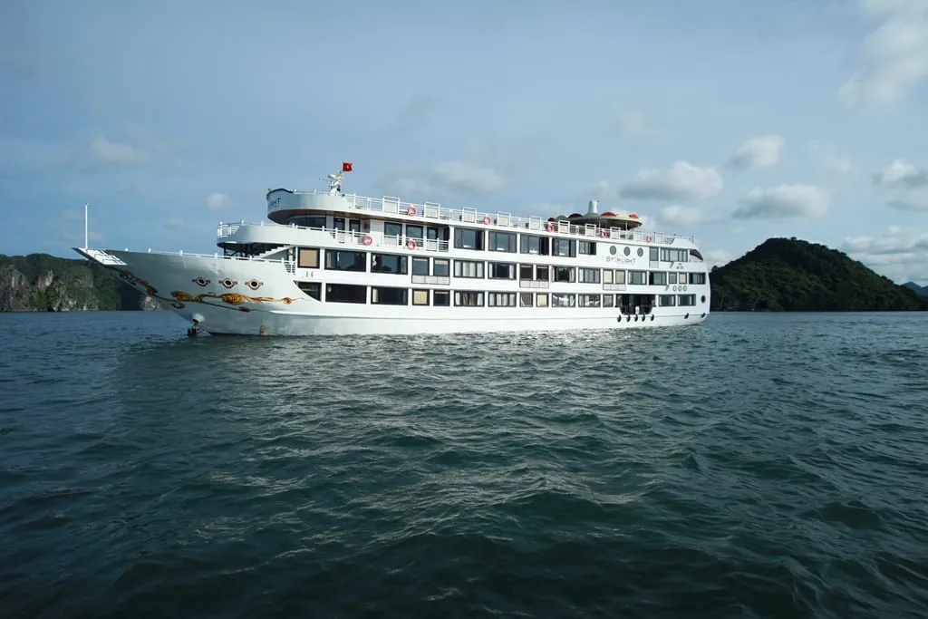 A large white multi-deck cruise ship with a golden dragon design on the bow and a Vietnamese flag, with 'Starlight' lettering on its side, sits on dark green water in front of forested limestone islands under a partly cloudy sky.