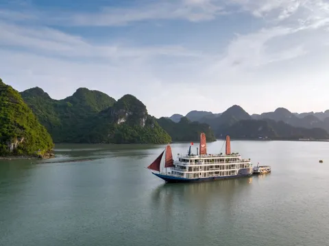 A multi-deck white cruise boat with a blue hull and red-orange sails sits on calm green water, with a small tender boat beside it and forested limestone karst islands in the background under a soft blue sky.