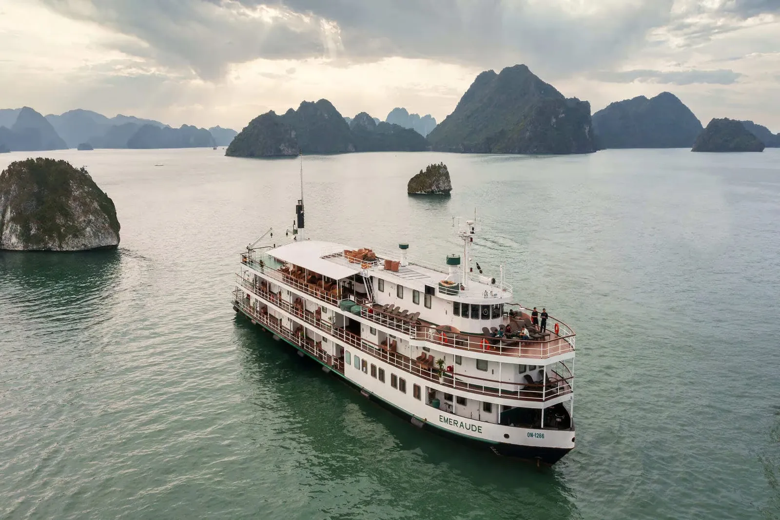 Aerial view of a white multi-deck cruise ship with 'EMERAUDE' on its stern, anchored in green water among limestone karst islands under a cloudy sky.