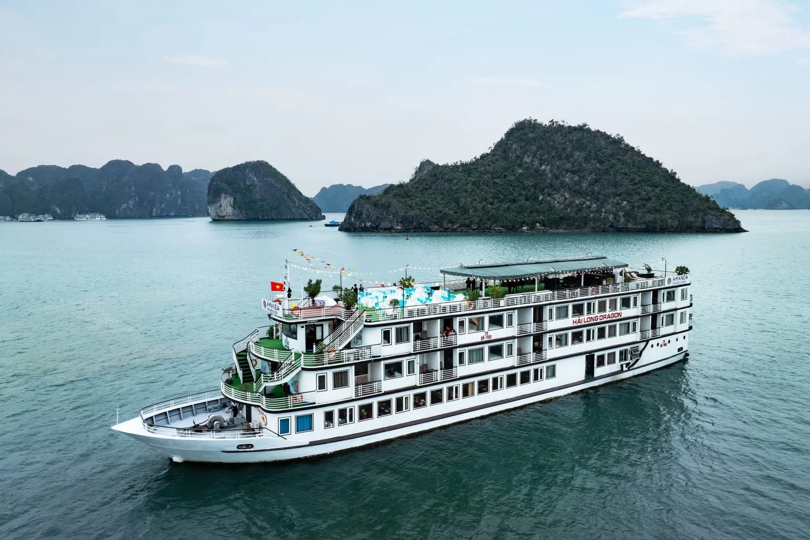 A white multi-deck cruise ship with green accents sails in calm turquoise water with limestone karst islands in the background.