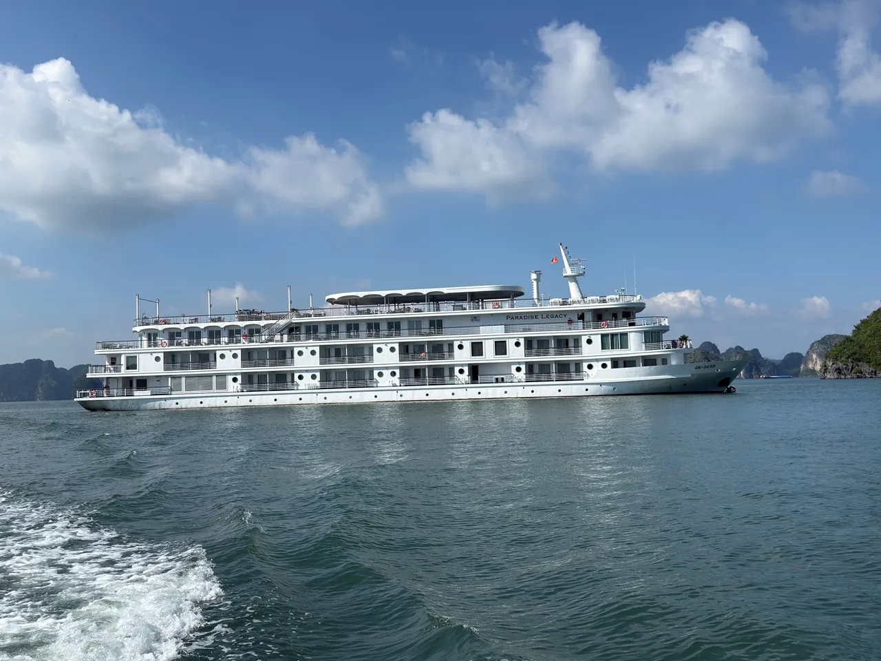 A large white multi-deck cruise ship with 'PARADISE LEGACY' lettered on its upper hull and QN-9698 on the bow, sailing on green water with limestone karst islands behind.