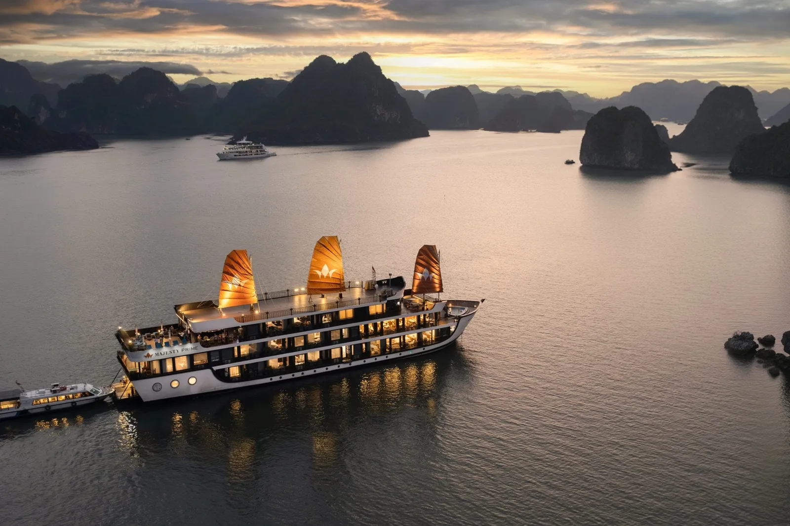 A traditional junk-style cruise ship with three orange sails illuminated at dusk floats among limestone karsts in calm bay waters.