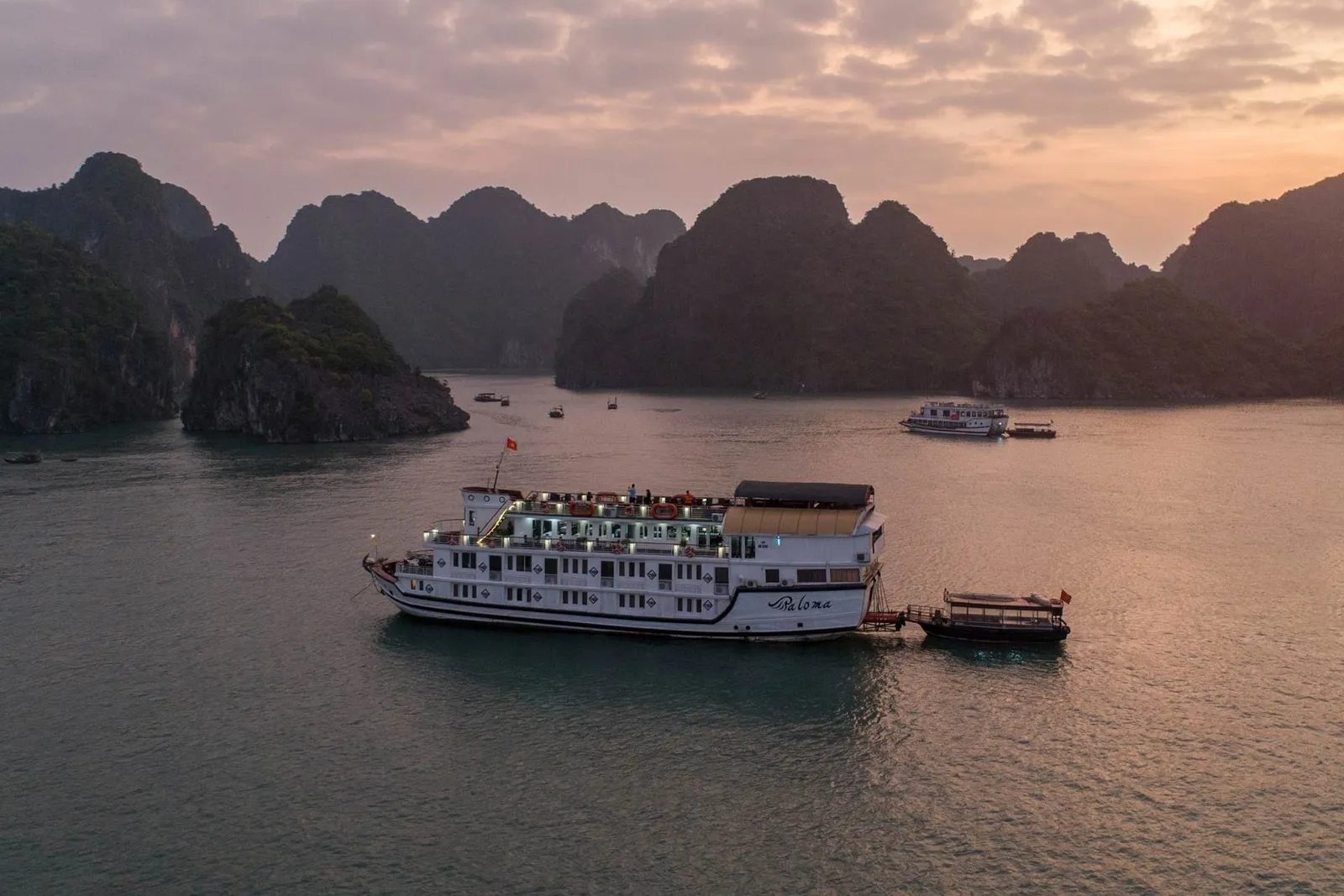 A white cruise boat with dark text on its hull floats among limestone karsts at sunset in calm water.