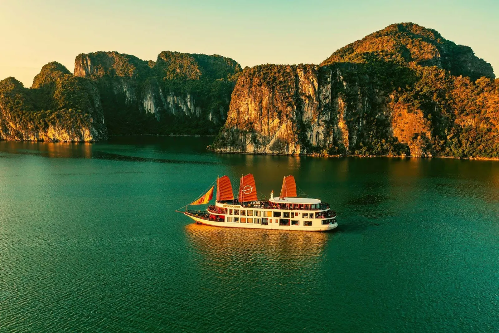 An aerial view of a white multi-deck cruise boat with orange-red sails and a circular logo on the center sail, floating on green water in front of steep forested limestone cliffs lit by golden light.