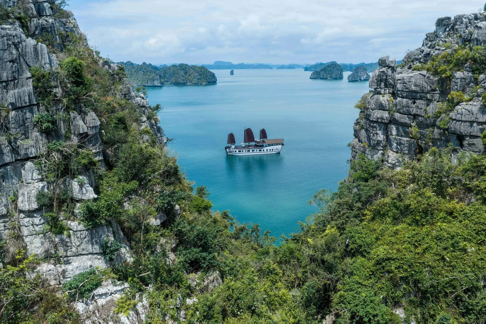 A white cruise boat with dark red sails sits on calm turquoise water, framed by grey limestone cliffs and green vegetation, with karst islands in the distance.