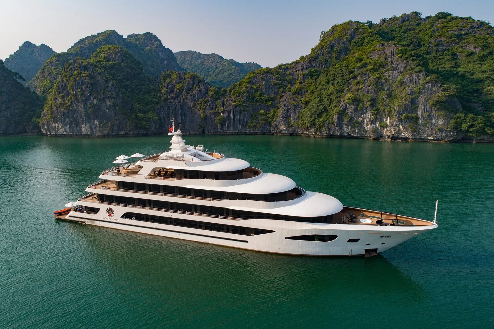 Aerial view of a white multi-deck luxury cruise ship with a lotus logo on the hull, anchored on green water in front of forested limestone karsts.