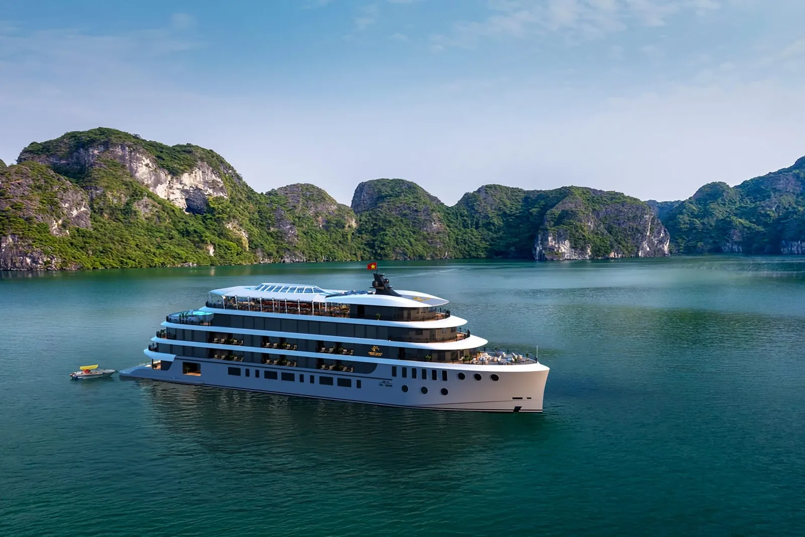 A large white multi-deck cruise ship with a small emblem on its hull and a Vietnamese flag sits on calm green water in front of forested limestone karst hills, with a small boat beside it.