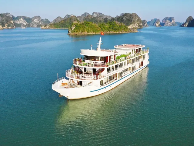 Aerial view of a large white multi-deck cruise boat with a red flag anchored on calm green-blue water, with forested limestone karst islands in the background.