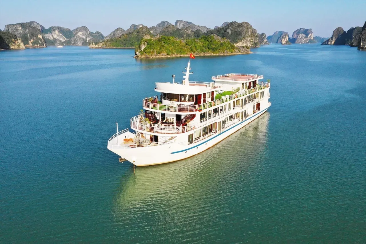 Aerial view of a large white multi-deck cruise boat with a red flag anchored on calm green-blue water, with forested limestone karst islands in the background.