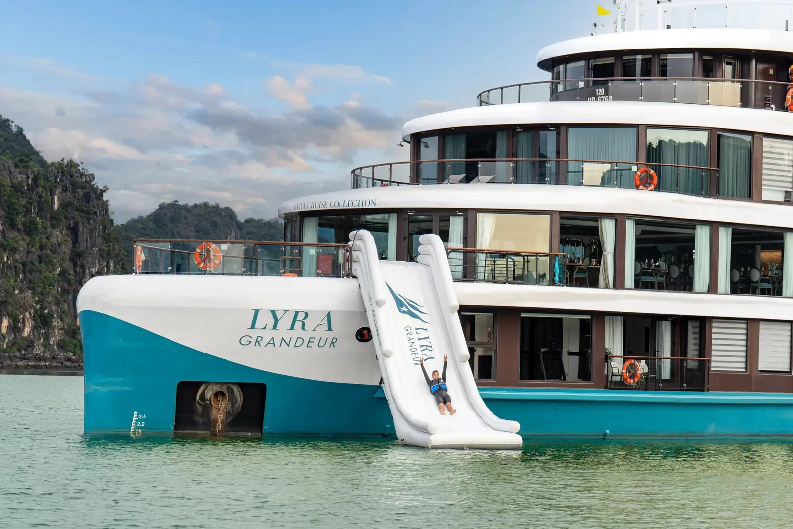 A white and turquoise cruise ship named LYRA GRANDEUR with an inflatable water slide deployed, anchored in calm green water with limestone karsts in the background.