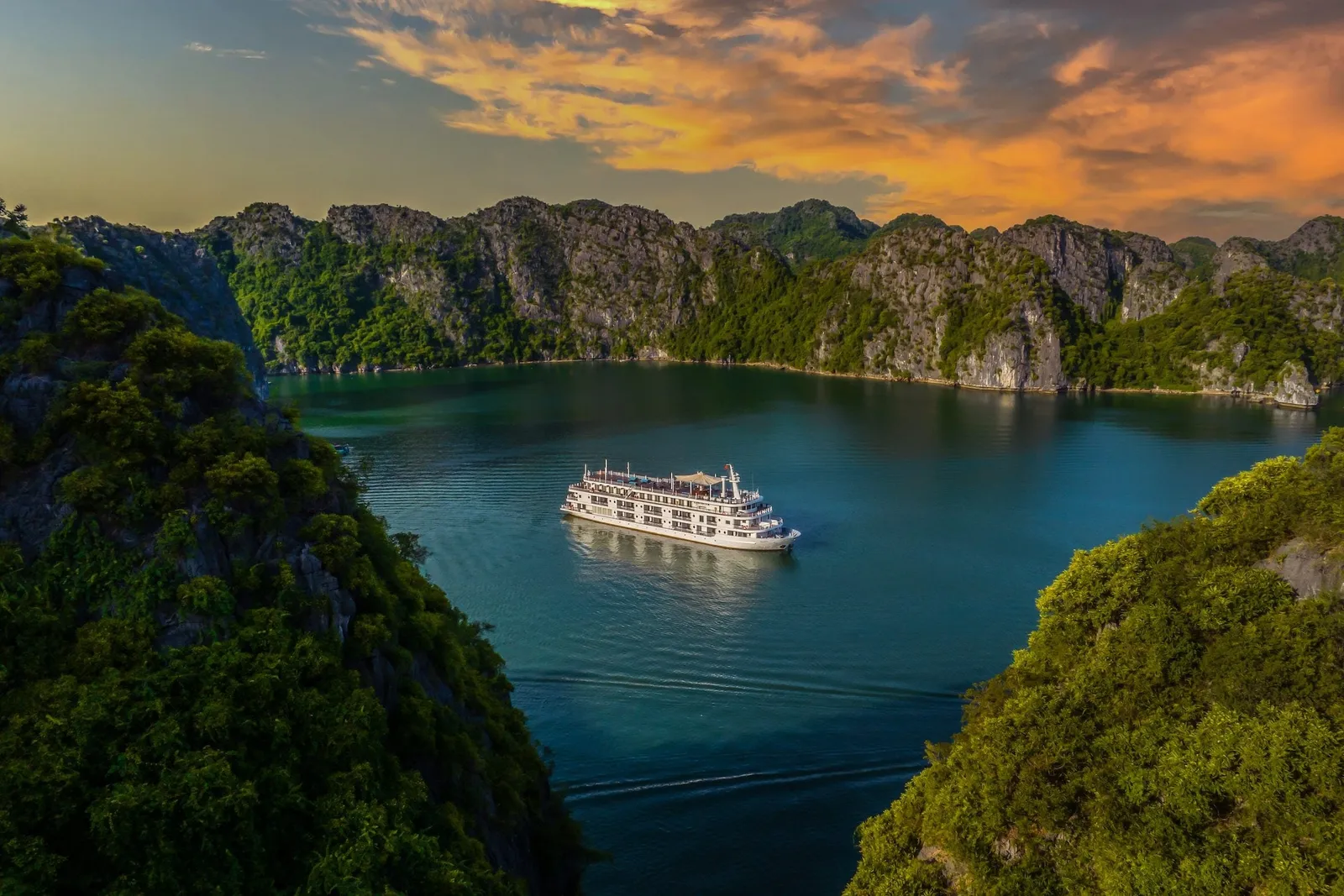 A white multi-deck cruise ship sits on calm turquoise water between forested limestone karst cliffs under an orange sunset sky.