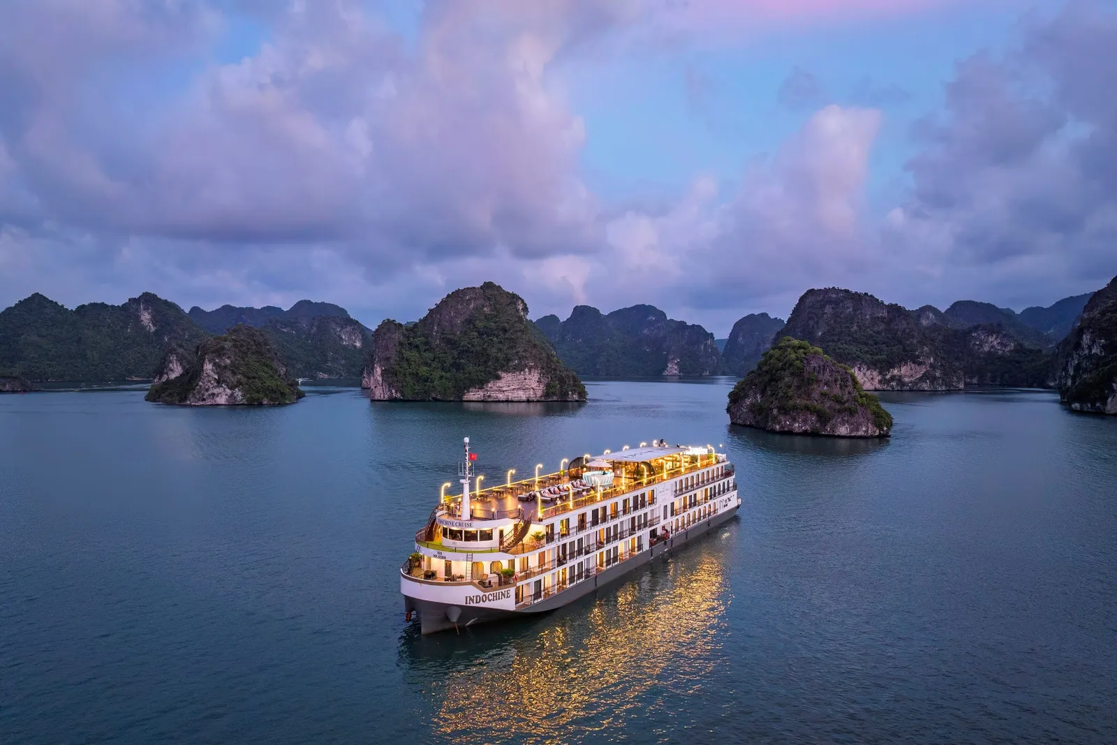 Indochine alone on still water under a violet sky, karst islands ranged along the horizon