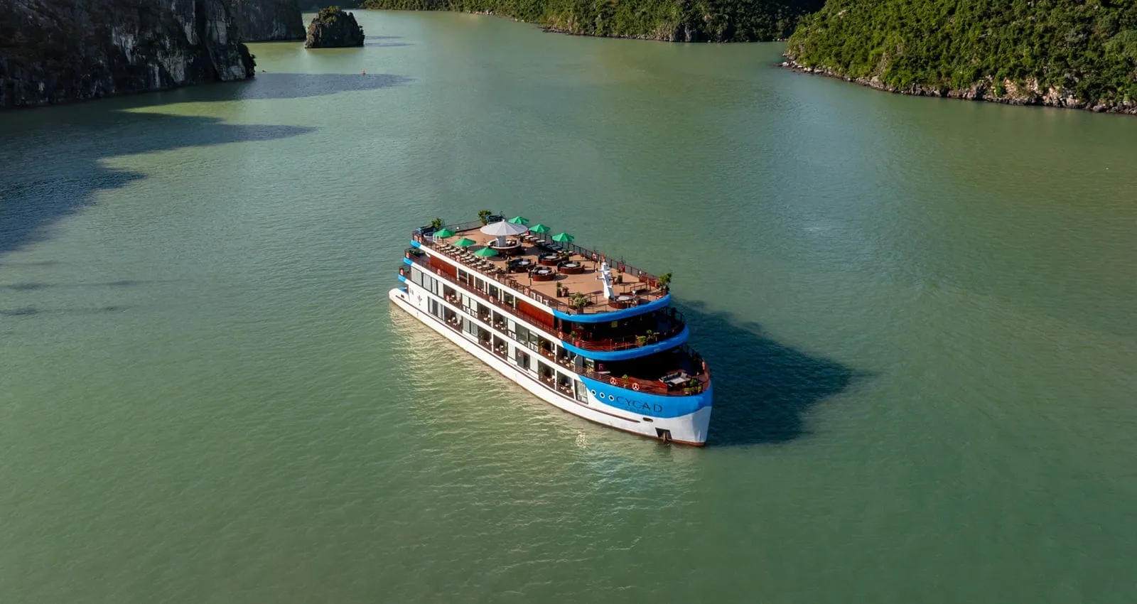 Aerial view of a white and blue multi-deck cruise ship with a rooftop deck of tables and green umbrellas, sitting in calm green water between forested limestone cliffs.