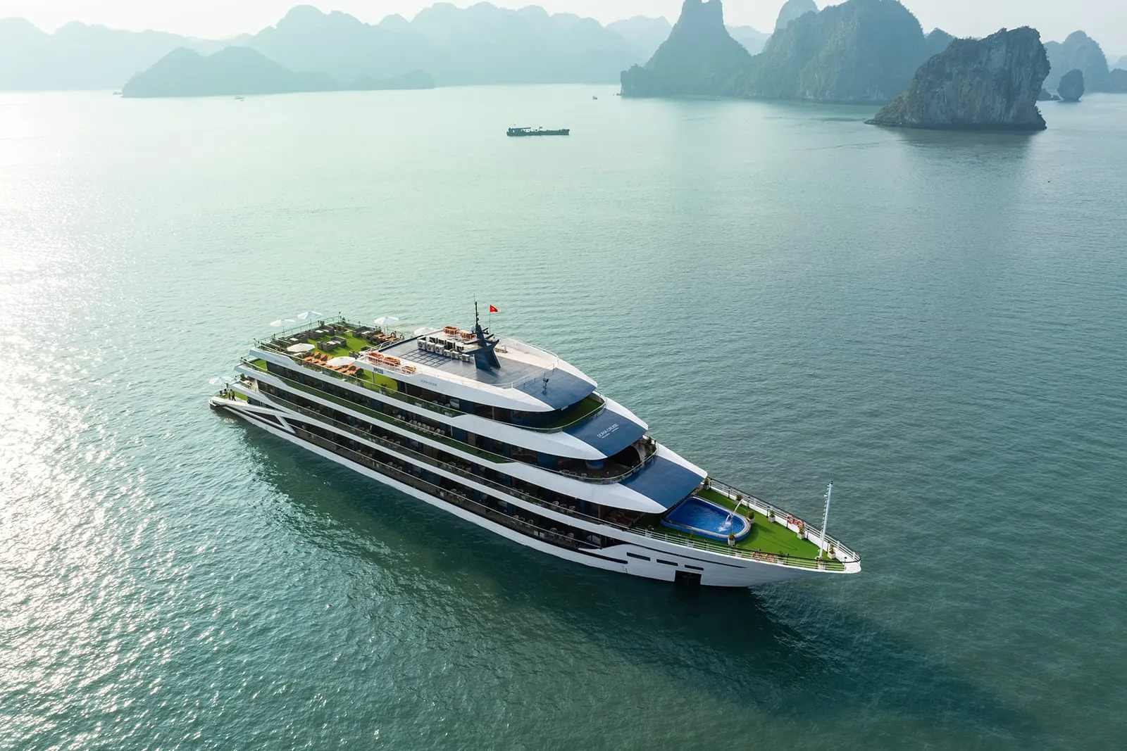A modern multi-deck cruise ship with white and blue hull sails in calm turquoise water surrounded by dramatic limestone karst islands.