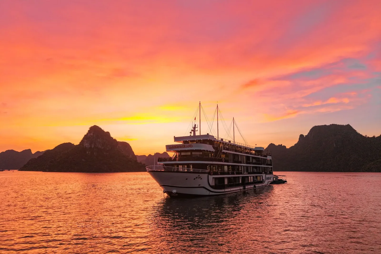 A multi-deck white cruise ship with name lettering on its upper superstructure floats on calm water at sunset, with a vivid orange and pink sky and dark limestone karst islands behind it.