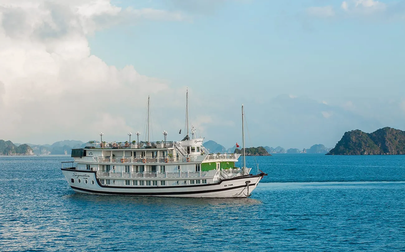 A multi-deck white cruise vessel with the name painted on its bow and a registration number on its stern, sailing on turquoise water with karst limestone islands in the background.