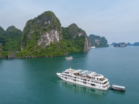 An aerial view of a white multi-deck cruise ship anchored in green-blue water surrounded by tall limestone karst islands.