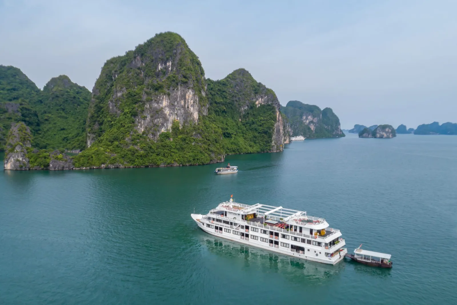 An aerial view of a white multi-deck cruise ship anchored in green-blue water surrounded by tall limestone karst islands.
