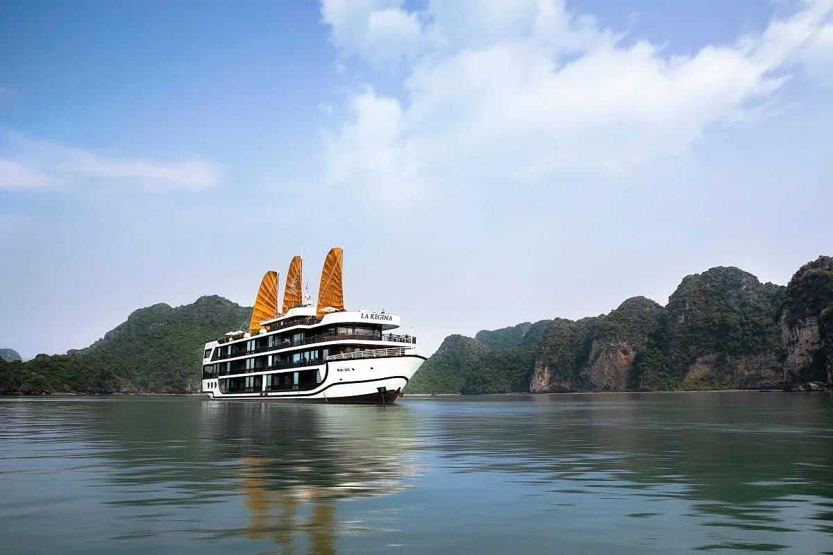 A white multi-deck cruise ship with orange junk-style sails and the name 'LA REGINA' on its upper structure floats on calm green water in front of forested limestone karst hills under a blue, partly cloudy sky.