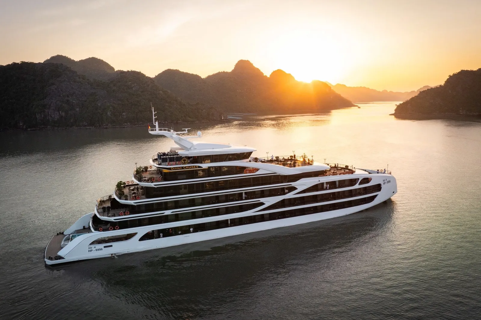 Aerial view of a large white multi-deck steel cruise ship sailing on calm water at sunset, with limestone karst islands silhouetted behind it.