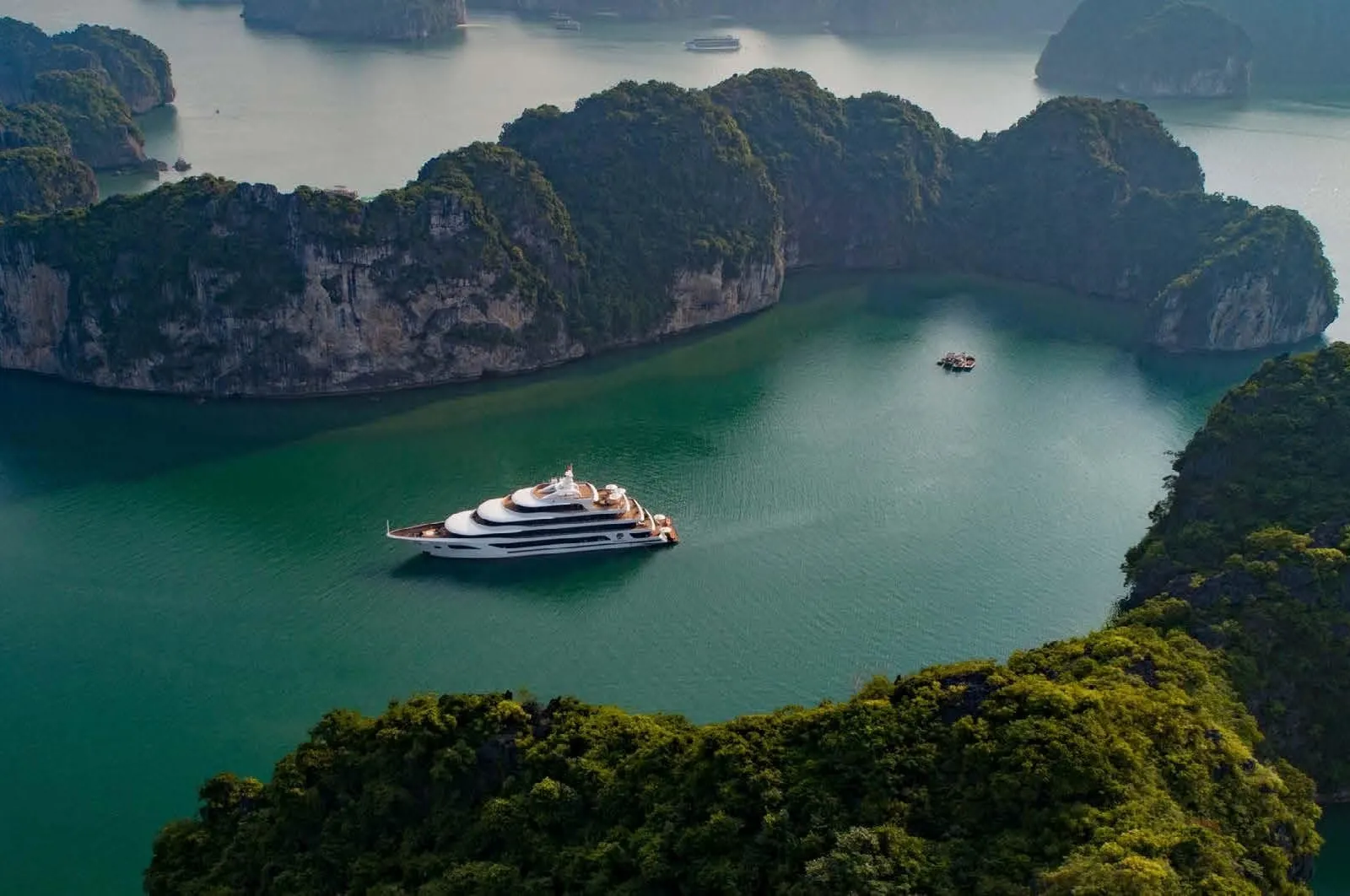 Aerial view of a white multi-deck cruise ship anchored in green water surrounded by forested limestone karst islands, with a small boat in the distance.