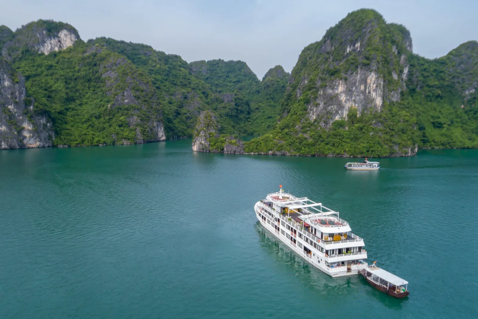 A white multi-deck cruise ship with a tender boat attached floats in emerald green water surrounded by tall limestone karst formations covered in vegetation.
