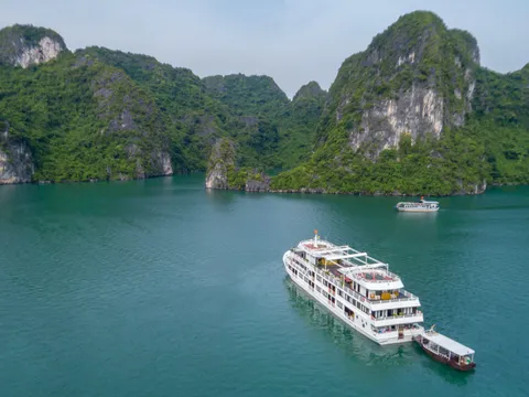 A white multi-deck cruise ship with a tender boat attached floats in emerald green water surrounded by tall limestone karst formations covered in vegetation.