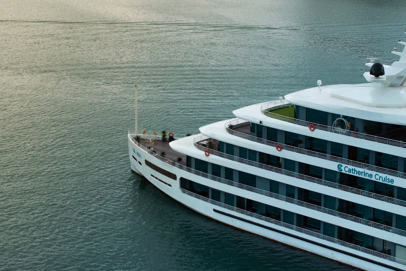Aerial view of the bow and upper decks of a white multi-deck steel cruise ship on calm green-grey water, with 'Catherine Cruise' written on its side.