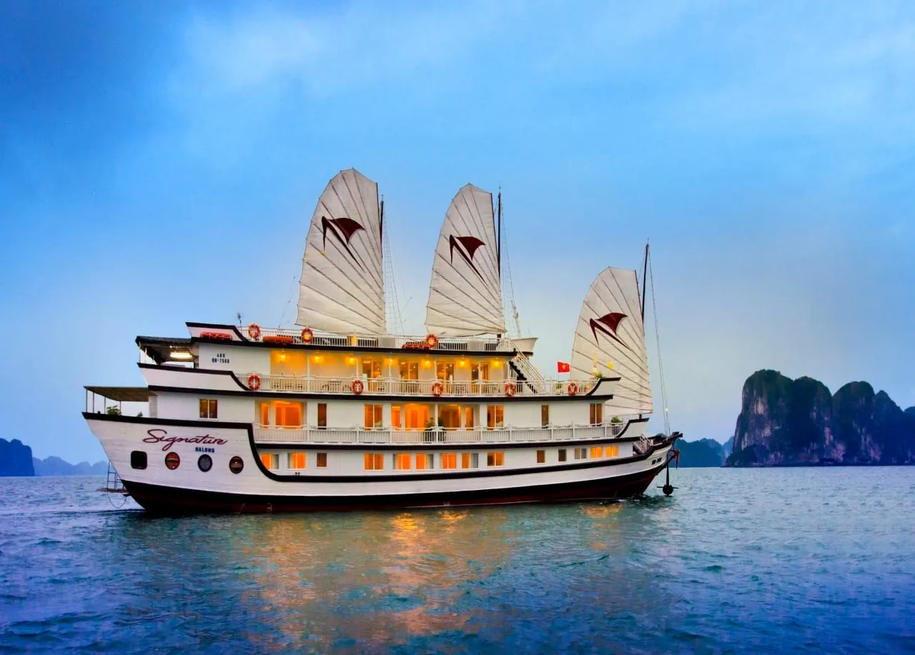 A white multi-deck junk cruise ship named 'Signature Halong' painted on its hull, with white sails, glowing cabin windows, and karst limestone formations visible in the background at dusk.