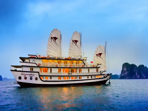 A white multi-deck junk cruise ship named 'Signature Halong' painted on its hull, with white sails, glowing cabin windows, and karst limestone formations visible in the background at dusk.