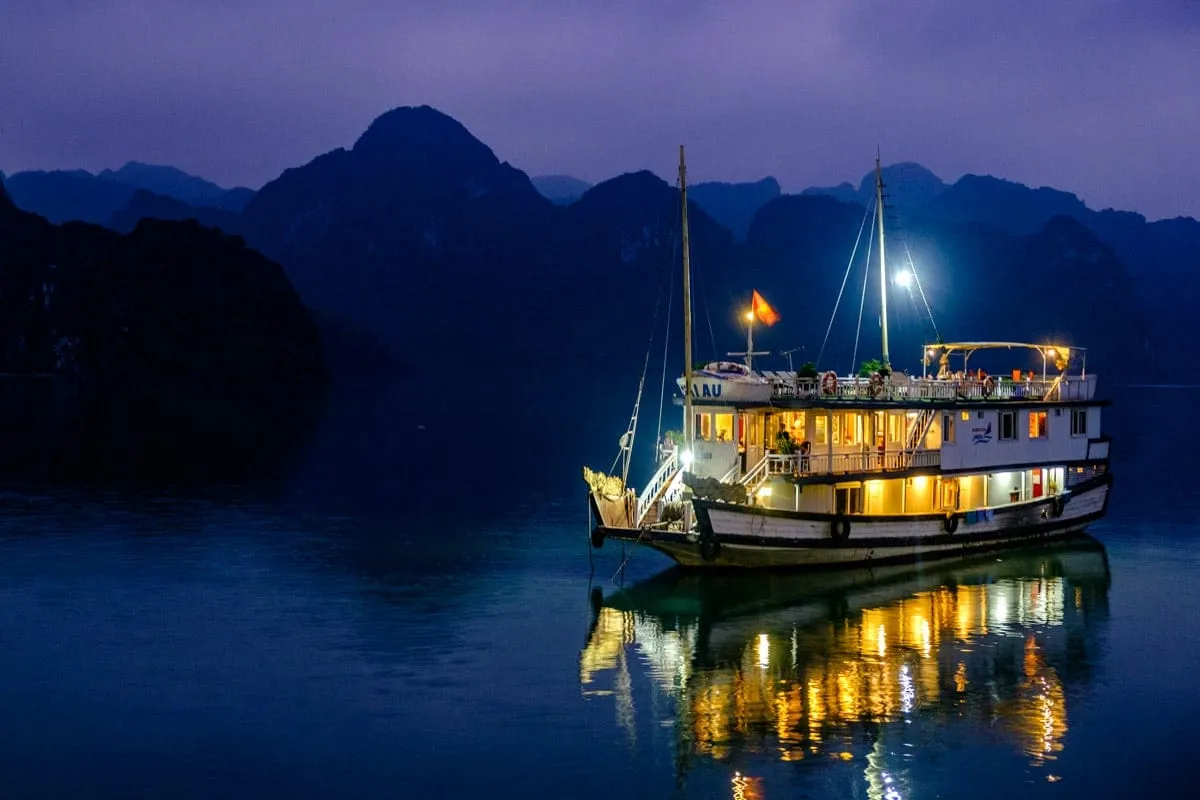 A white wooden cruise boat with yellow-lit windows and a Vietnamese flag anchored in calm water at dusk with dramatic limestone karst mountains silhouetted in the background.