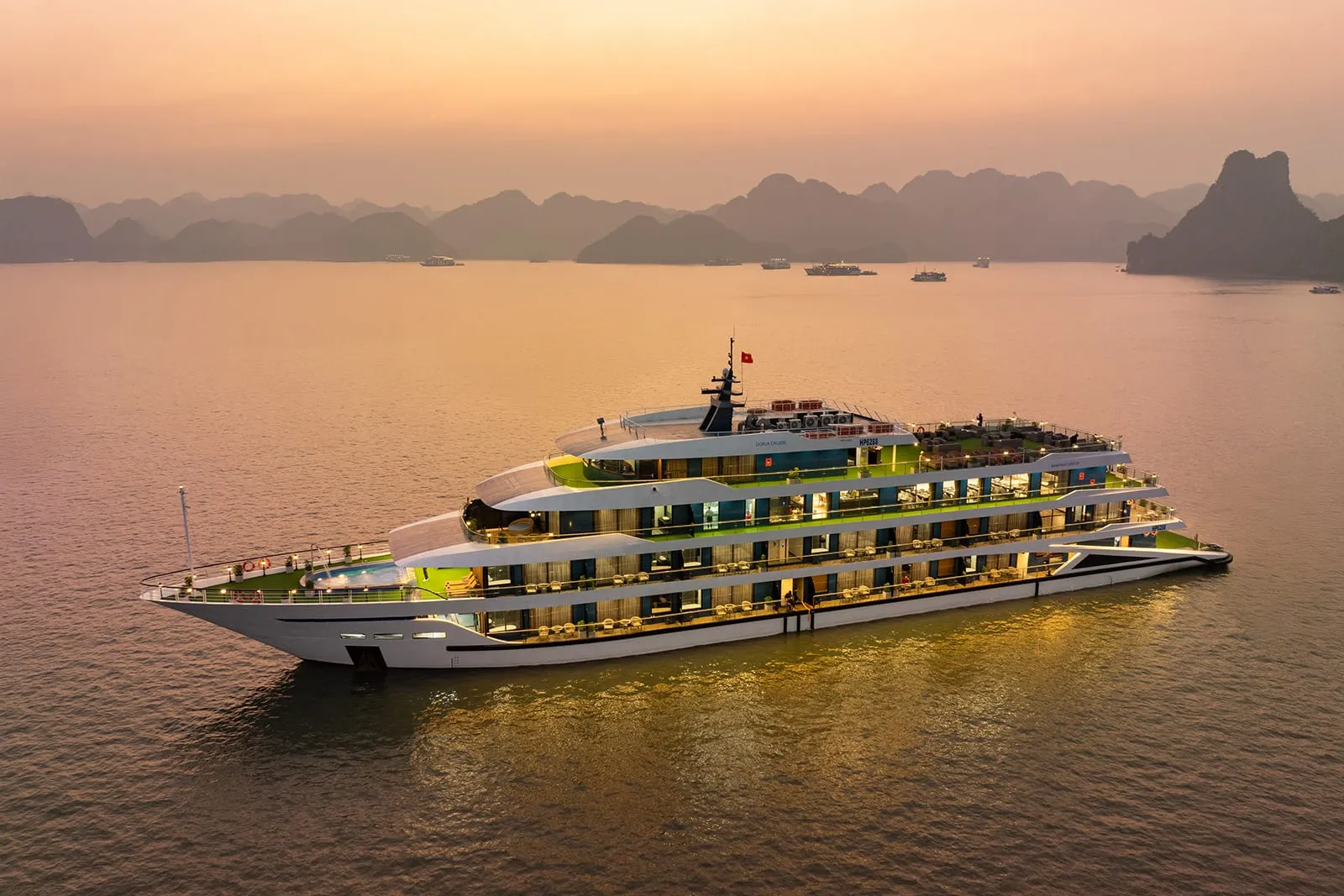 A modern multi-deck cruise ship with bright green upper decks and white hull sails through calm waters at sunset with karst limestone mountains in the background.