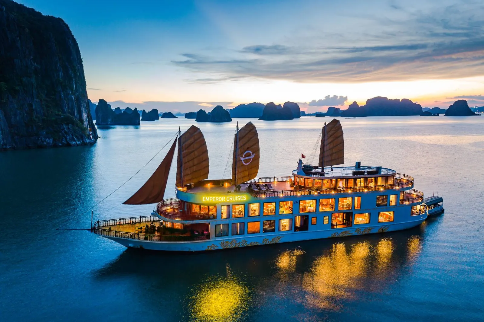 An aerial dusk view of a lit-up multi-deck junk-style cruise boat with brown sails and an illuminated 'Emperor Cruises' sign, anchored among limestone karst islands under a colorful sunset sky.