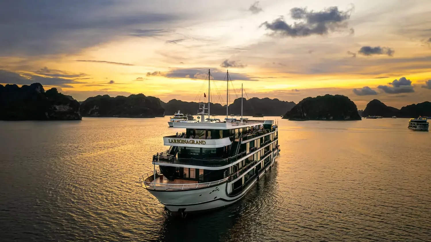 An aerial view of a multi-deck cruise ship with 'LA REGINA GRAND' lettered on its upper structure, anchored among dark limestone islands under a golden sunset sky.