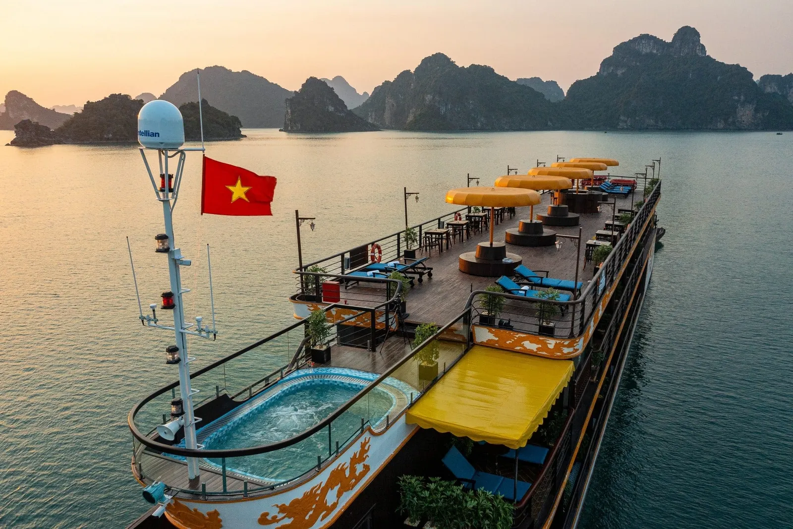 Aerial view at sunset of a multi-deck cruise ship's sun deck with a plunge pool, yellow umbrellas, sun loungers and a Vietnamese flag, surrounded by limestone karsts.