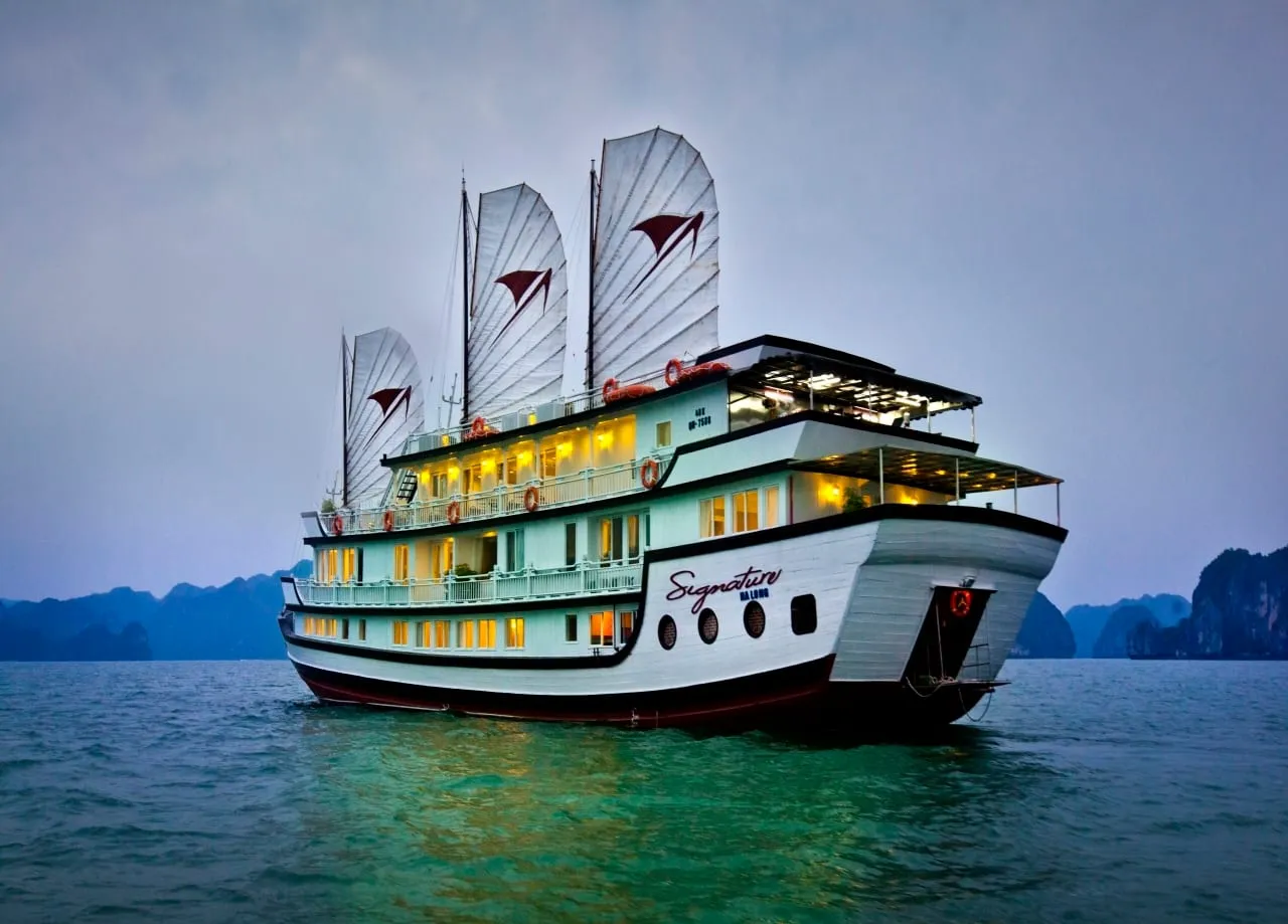 A multi-deck white junk cruise ship named 'Signature Ha Long' painted on its hull, lit with warm interior lights, sailing on green-blue water with karst limestone islands in the background at dusk.