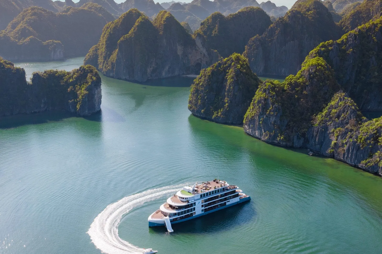 Aerial view of a white and blue multi-deck cruise ship in green bay water among karst limestone islands, with a small speedboat leaving a curved white wake beside it.