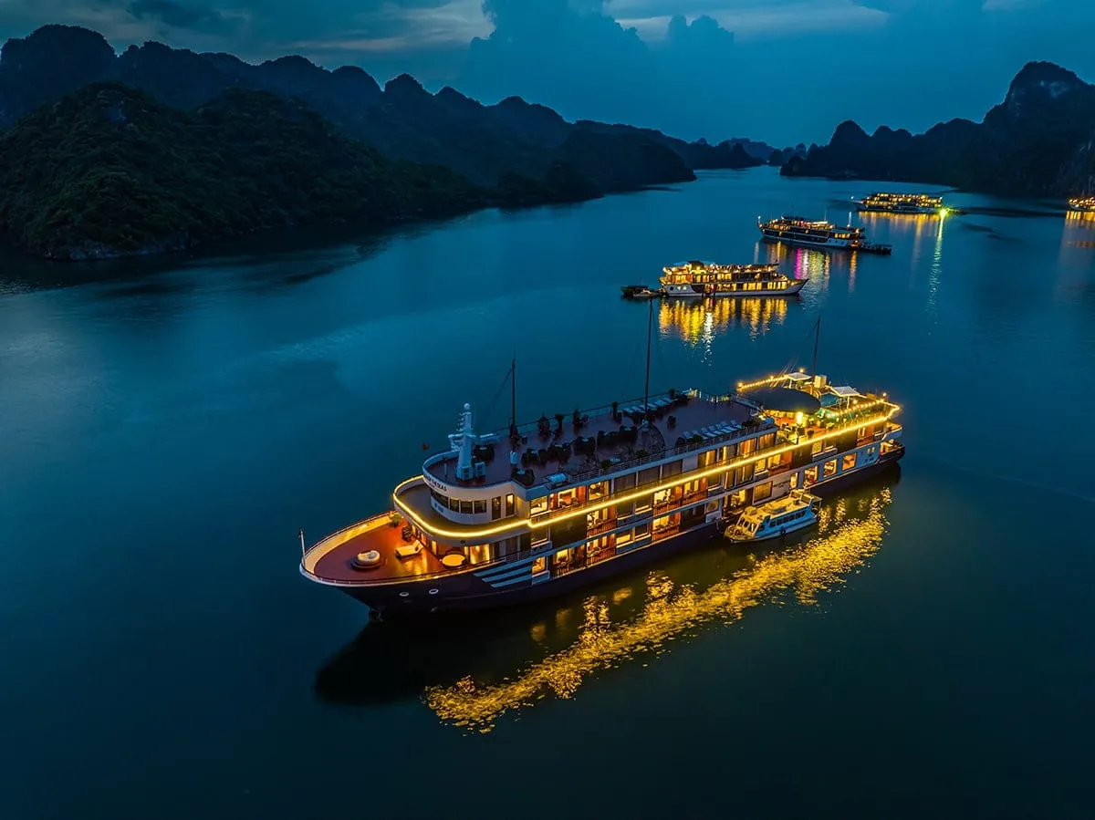 Aerial dusk view of a lit-up multi-deck steel cruise ship anchored among karst islands, with other illuminated cruise boats in the background and golden reflections on calm water.