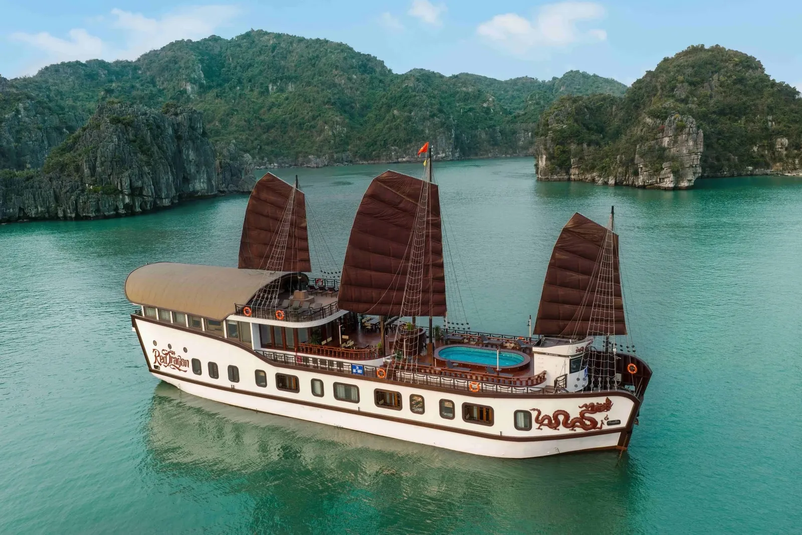 An aerial view of a white junk-style cruise boat with brown sails, a small pool on the upper deck, and a dragon painted on the hull, with 'Red Dragon' lettering on the side, floating on green water among forested limestone karst islands.