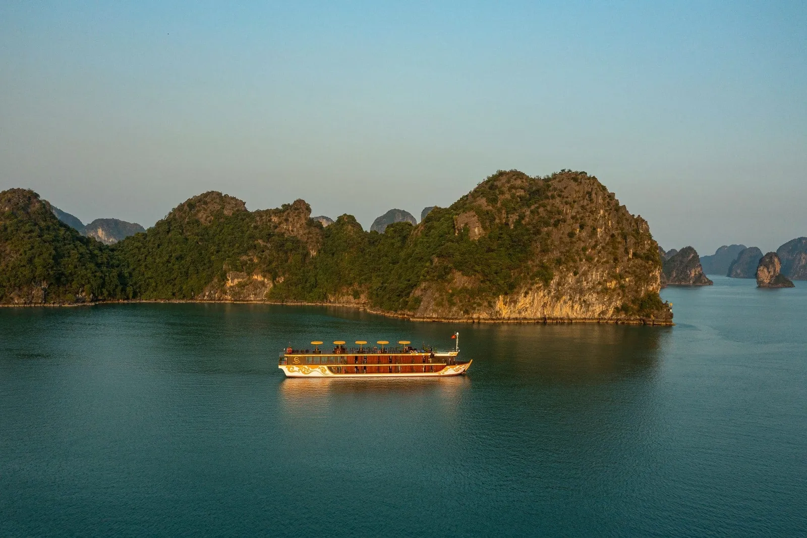 Aerial view of a two-deck wooden-style boat with a white hull, gold decorative motifs and small yellow umbrellas on the top deck, cruising on calm green water beside forested limestone karst islands at golden hour.