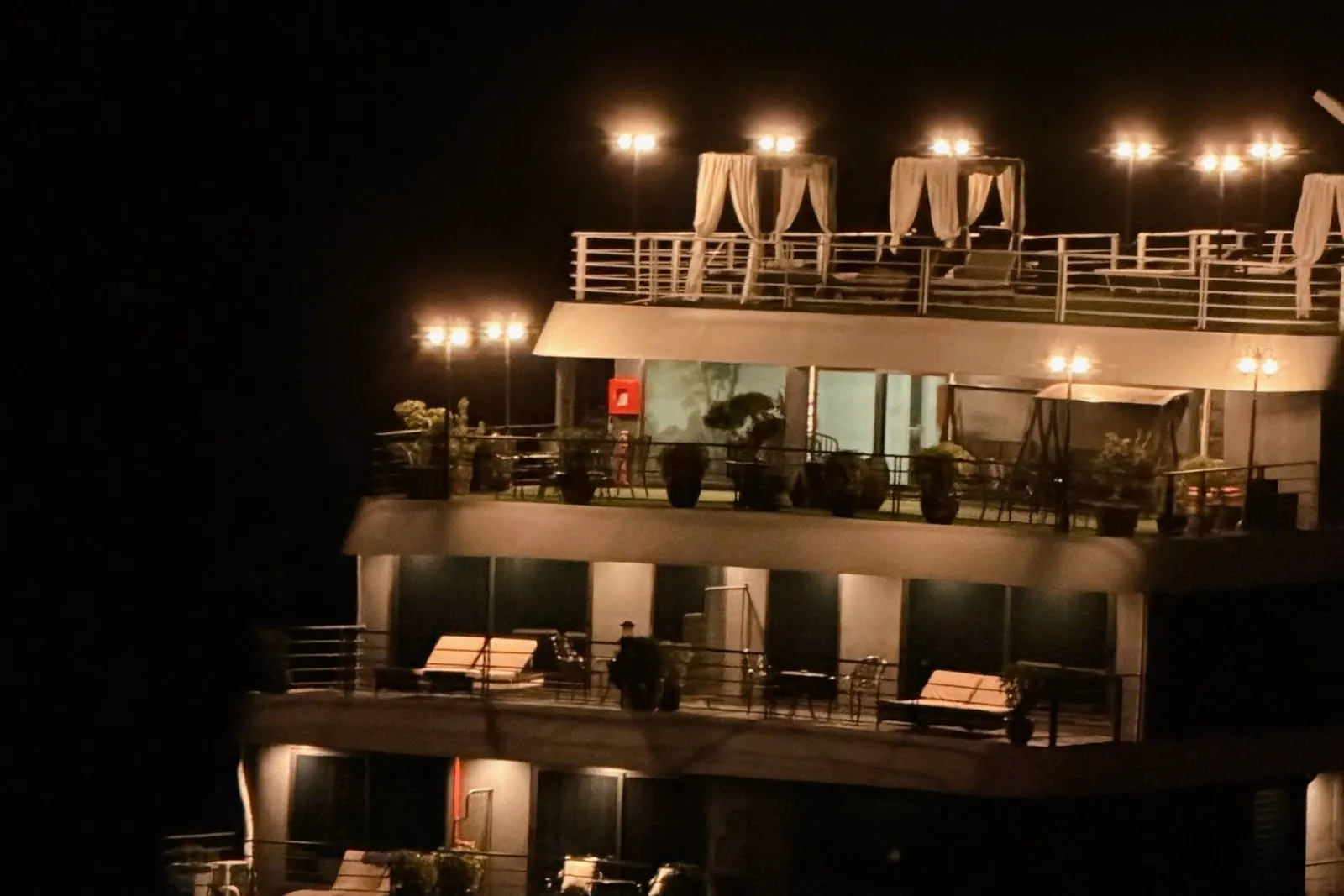 A multi-deck steel cruise ship lit up at night, with balconies, loungers, white-curtained canopies on the top deck and a small pool on the lower deck, seen from outside against a dark sky.