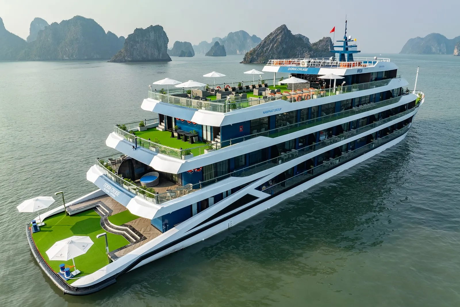 A large multi-deck white and navy cruise ship with bright green artificial turf decks, white umbrellas, and passengers aboard floats in calm water surrounded by limestone karst islands.