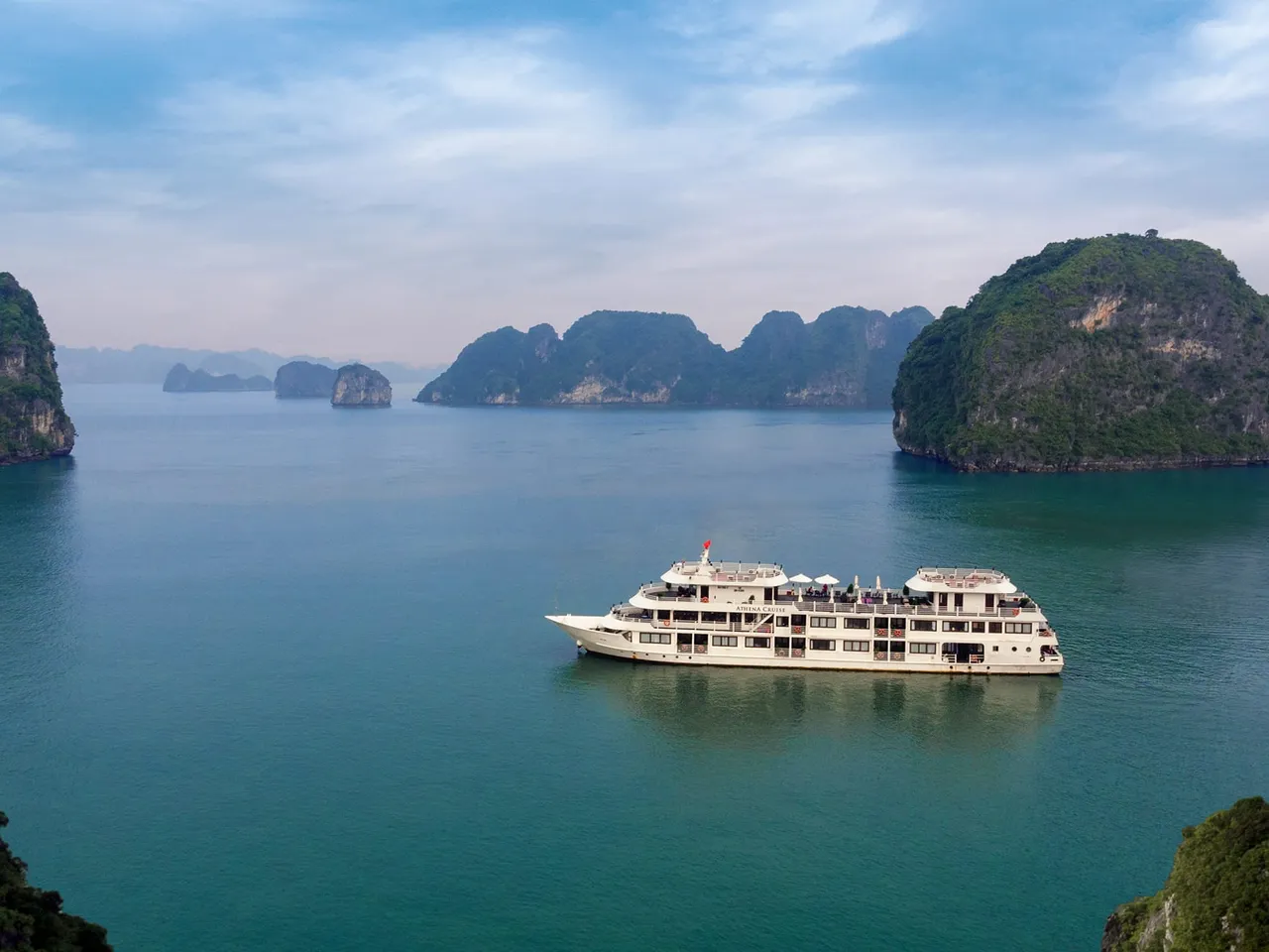 A white multi-deck cruise ship floats in calm turquoise water surrounded by dramatic limestone karst islands.
