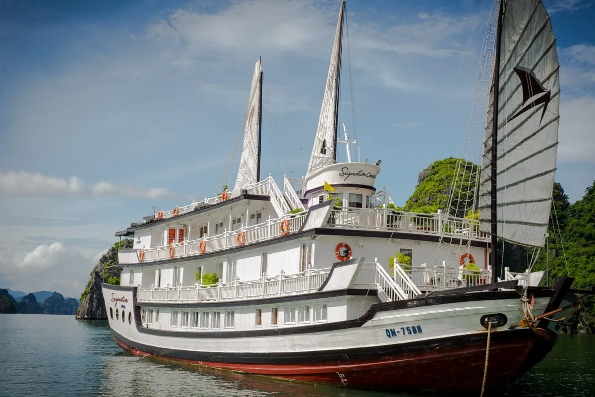 A white multi-deck junk-style cruise vessel with dark sails, registration number QN-7588 and the name 'Signature' painted on the hull, anchored among karst limestone islands.