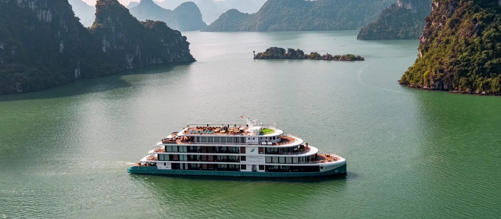 Aerial view of a white and teal multi-deck cruise ship on green water among limestone karst islands.