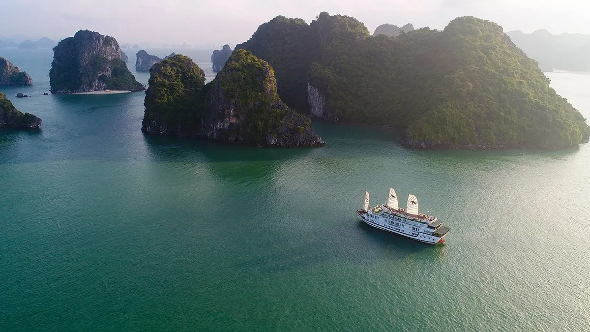 An aerial drone shot of a multi-deck cruise junk with white sails sailing on green water surrounded by karst limestone islands.